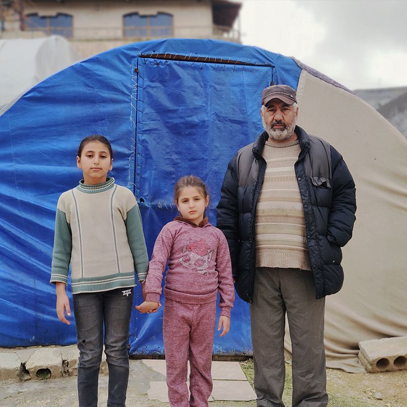 A father standing with his two daughters outside the tent that he's been living in with his family since the Turkey-Syria earthquakes.