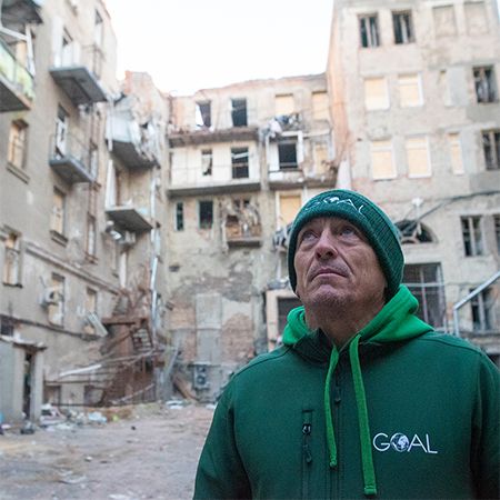 A man wearing a green hoodie and hat with GOAL logo standing in front of buildings in Ukraine that have been damaged by the conflict with Russia