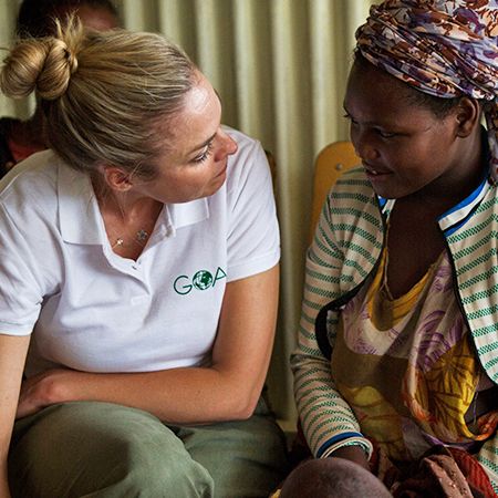 Two women speaking to each other. Woman on the left is wearing a white top with GOAL logo and woman on the left is a happy beneficiary