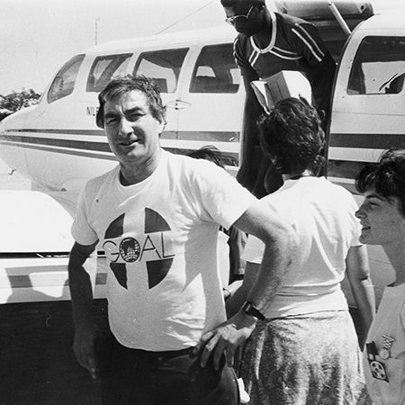 A man, John O'Shea founder of GOAL, standing in front of a plane wearing GOAL t-shirt in 1977