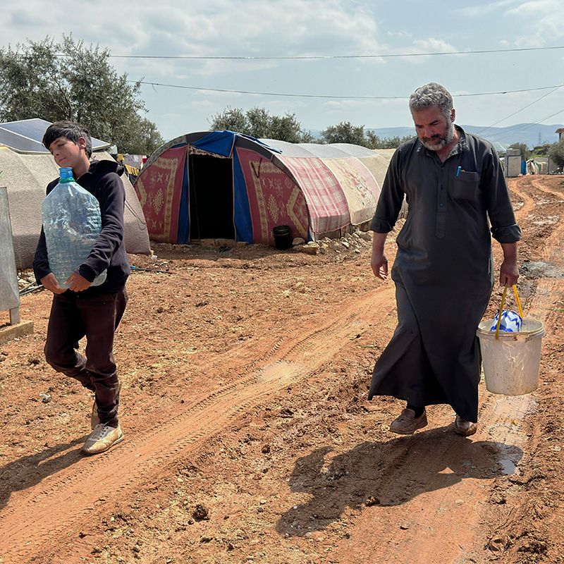 A teenage boy on the left carrying a large gallon of water with his father on the right carrying a bucket of water back to their tent in displacement camp in Idleb, Northwest Syria