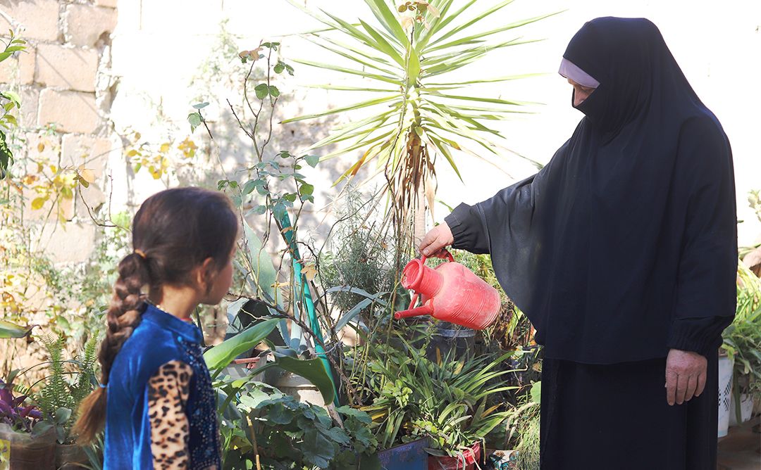 SANAD Cycle 2_SY_Amina Family_Garden_body A woman beneficiary of GOAL standing on the right watering her garden while her granddaughter looks on from her left side.